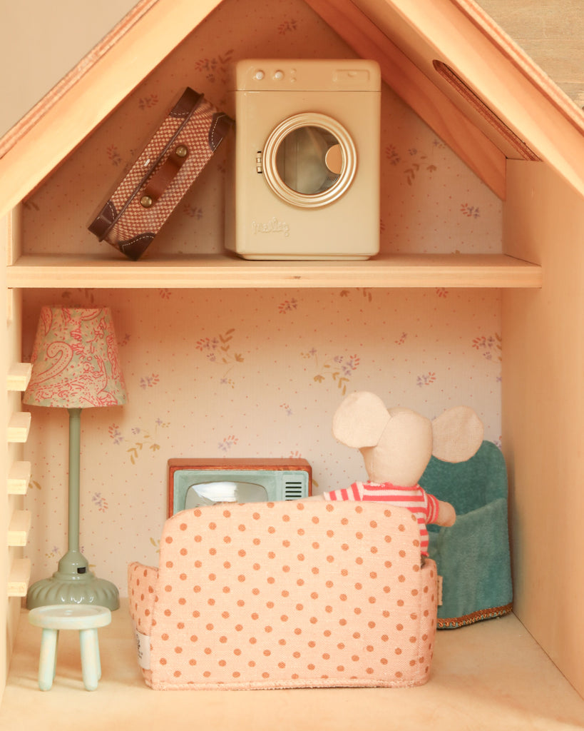 Experience the charm of the Maileg Fully Furnished Farmhouse Set + Surprise Gift, featuring a cozy room with a mouse doll relaxing on a pink polka dot sofa, surrounded by adorable dollhouse accessories—including a miniature TV, lamp, washing machine, and suitcase—all beautifully arranged against pastel wallpaper.