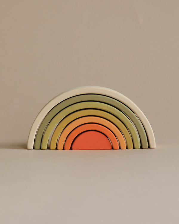 A set of colorful wooden nesting arches forming a rainbow, arranged from largest to smallest with non-toxic paint in muted colors including green, yellow, and orange, against a plain beige background.
