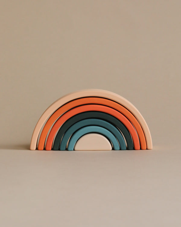 A stack of Handmade Mini Rainbow Stackers in shades of teal, blue, orange, and peach, arranged against a light beige background. This stacking toy encourages open-ended play.