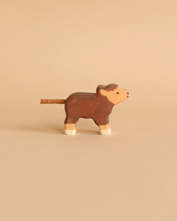 A small wooden toy cow with a rope tail is depicted against a beige background. Made in Europe, the Holztiger Highland Cattle, Small figure is brown with white accents on its legs and face, featuring a simple, hand-painted design.