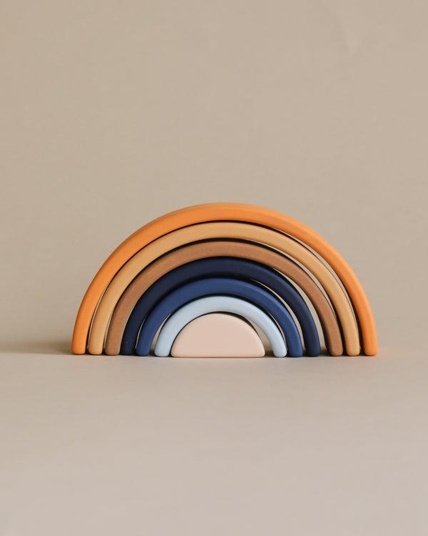 A set of Handmade Mini Rainbow Stackers - Desert Night, painted with non-toxic paint in a gradient of shades from orange to blue, neatly arranged in order of size against a soft beige background.