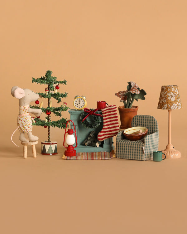 The Maileg Little Sister Cozy Christmas Living Room Set features a mouse decorating a mini Christmas tree beside a checked armchair, side table, clock, lantern, cup, floor lamp, and accessories, all on a warm beige background.