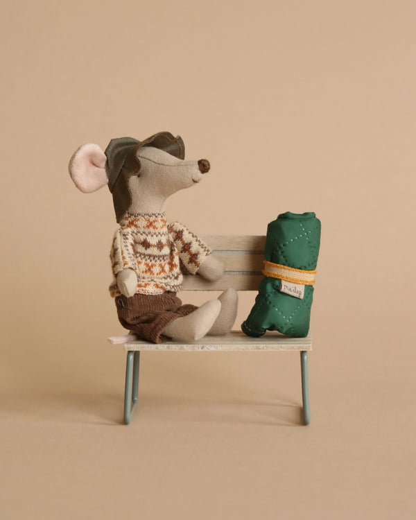 A Maileg Hiker Mouse, Big Brother dressed in a sweater and cap sitting on a metal bench beside a miniature green backpack. The background is a soft tan color.