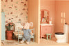 A Maileg Fully Furnished Farmhouse Set + Surprise Gift features a small plush mouse in striped clothes sitting on a toy bed in a charming farmhouse dollhouse room, surrounded by delightful accessories like a plant, wallpaper, vanity, and toilet.