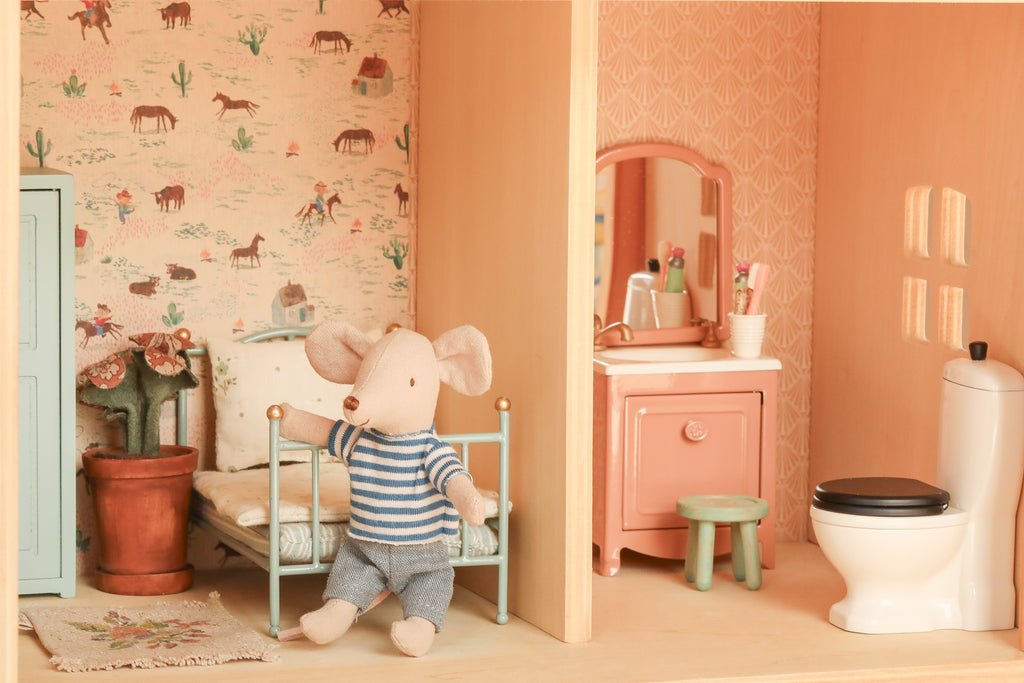 A Maileg Fully Furnished Farmhouse Set + Surprise Gift features a small plush mouse in striped clothes sitting on a toy bed in a charming farmhouse dollhouse room, surrounded by delightful accessories like a plant, wallpaper, vanity, and toilet.