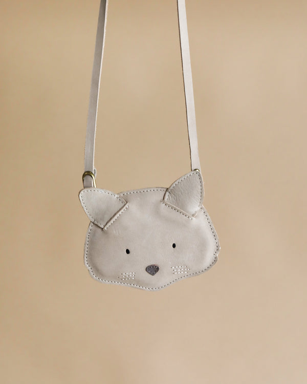 A small, light gray premium leather purse shaped like a cat's face, with pointy ears and facial features stitched in black, hanging from a thin strap against a beige background.