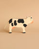 A handcrafted wooden toy cow with a simple, minimalist design stands on a beige background. Made from maple and beech wood, this Holztiger Black Cow, Standing is painted white with black spots, has a pink snout, and a rope tail. Its shape and features are stylized rather than realistic.