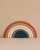 A stack of wooden rainbow arches in graduated sizes, each coated with non-toxic paint in different earthy colors ranging from deep teal to warm terracotta, arranged against a beige background.