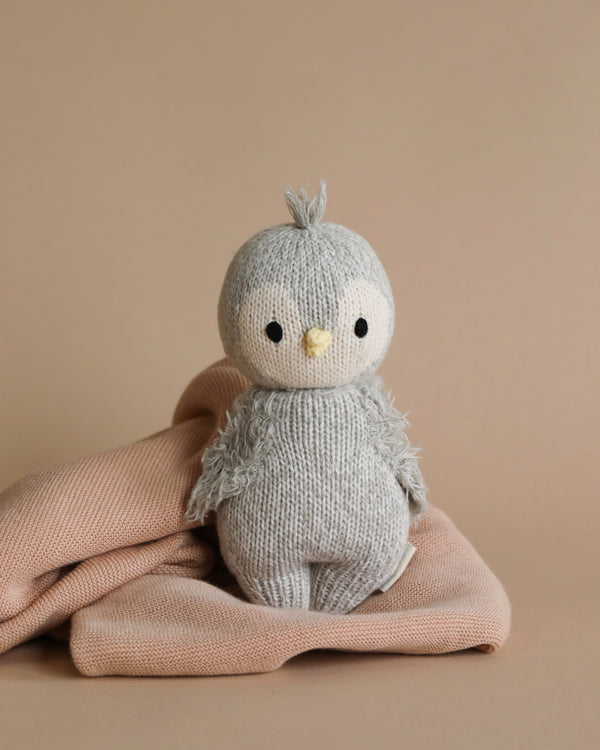 A cute, hand-knit Cuddle + Kind Baby Penguin toy bird with a tuft of hair on top, sitting on a pink fabric against a tan background.