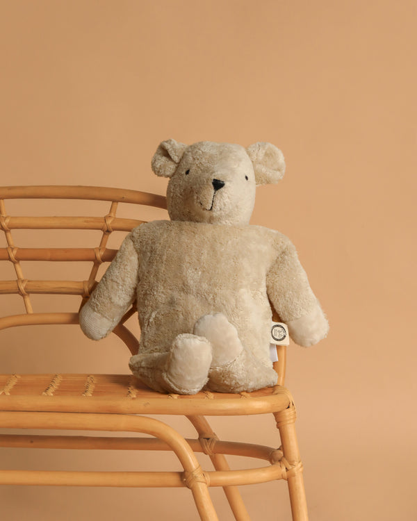 A Senger Naturwelt Cuddly Animal - Linen Bear sits upright on a light wooden chair against a plain beige background.