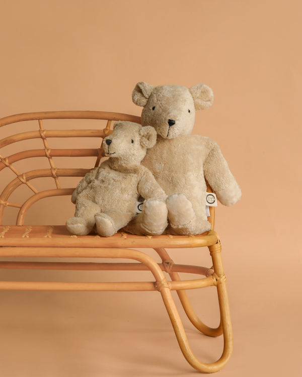 Two Senger Naturwelt Cuddly Animal - Linen Bears, one large and one small, sit side by side on a rattan bench against a plain beige background.