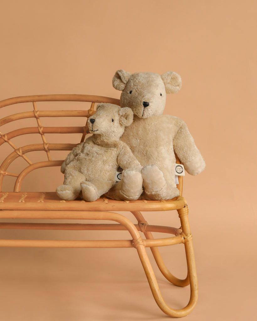 Two Senger Naturwelt Cuddly Animal - Linen Bears, one large and one small, sit side by side on a rattan bench against a plain beige background.