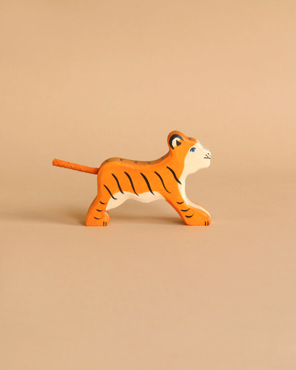 A small, orange, wooden Holztiger Tiger, Small, Running handcrafted from HOLZTIGER figures is walking towards the right side of the image. The light beige background complements the tiger's color. The toy, made in Europe, features black stripes, white accents, and blue eyes.