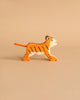 A small, orange, wooden Holztiger Tiger, Small, Running handcrafted from HOLZTIGER figures is walking towards the right side of the image. The light beige background complements the tiger's color. The toy, made in Europe, features black stripes, white accents, and blue eyes.