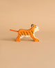 A handcrafted tiger from the Holztiger Handmade Wooden Safari Animals Set, featuring orange and black stripes, stands on a light beige surface with a matching background.
