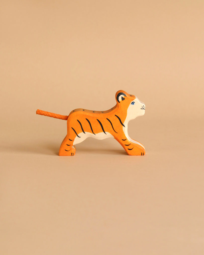 A handcrafted tiger from the Holztiger Handmade Wooden Safari Animals Set, featuring orange and black stripes, stands on a light beige surface with a matching background.