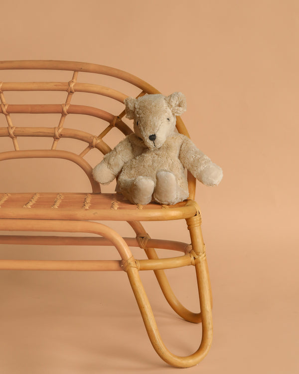 A Senger Naturwelt Cuddly Animal - Linen Bear sits on a light brown rattan bench against a plain beige background.