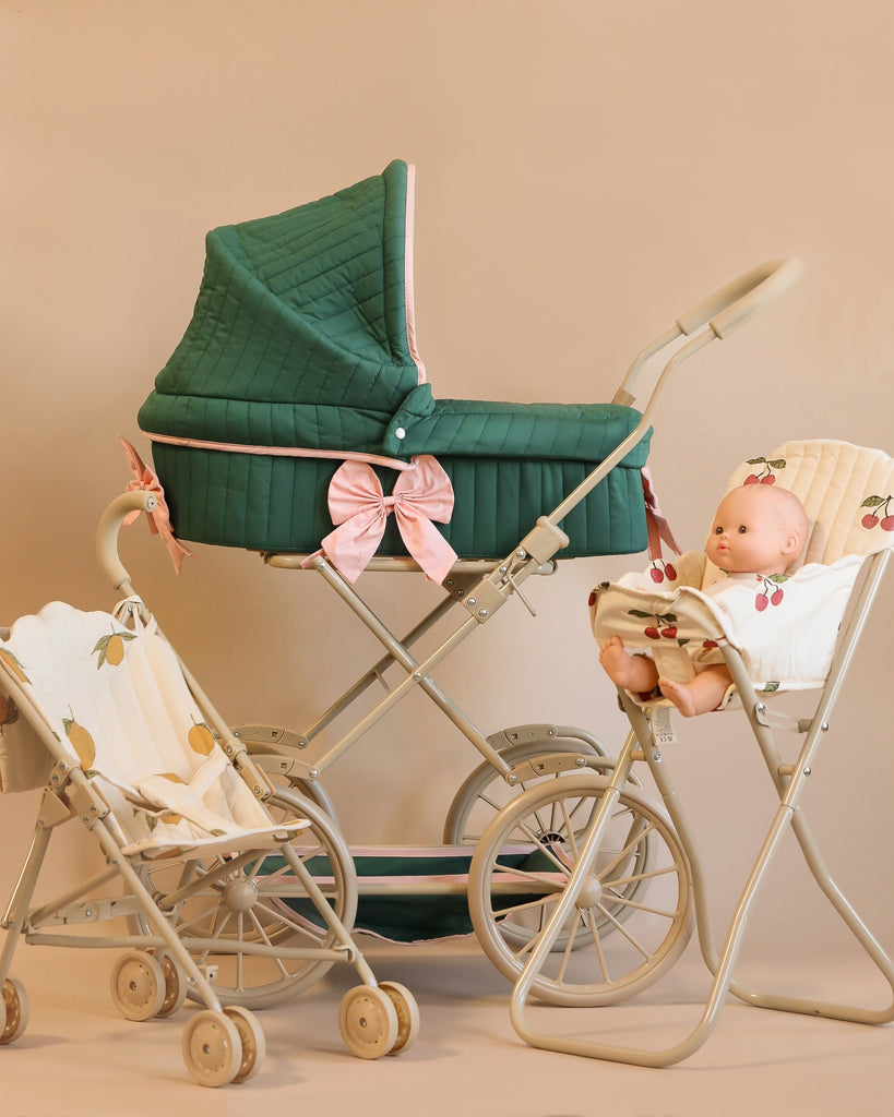 A vintage-style baby stroller in green, embellished with a pink bow, is accompanied by two smaller play prams crafted from recycled materials. One of the Doll Pram - Bowie Smoke Pine models carries a doll, and both are adorned with fruit-patterned fabric made from organic cotton, enhancing their charming and eco-friendly design.