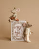 Two small, Royal Twins in Box fabric mouse figurines with crowns, one seated on a book and one standing beside it, against a beige background. The book features intricate illustrations of mice. One mouse is dressed in