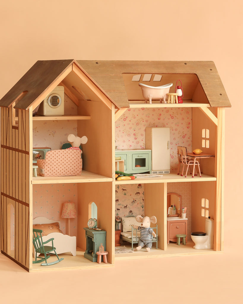 The Maileg Fully Furnished Farmhouse Set + Surprise Gift features a wooden farmhouse with six rooms, complete with charming Maileg Mouse Furniture and delightful Dollhouse Accessories such as beds, chairs, kitchen appliances, a bathtub, and a small stuffed animal seated in the living room. The scene is set against a soft peach background.