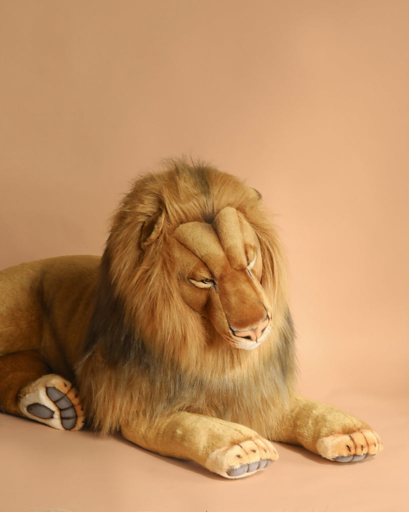 The Extra Large Lion, 39'' – FINAL SALE, is a realistic plush with hand-sewn details, lying on a light tan surface with its front paws stretched forward and its head turned slightly to the side against a matching tan background.