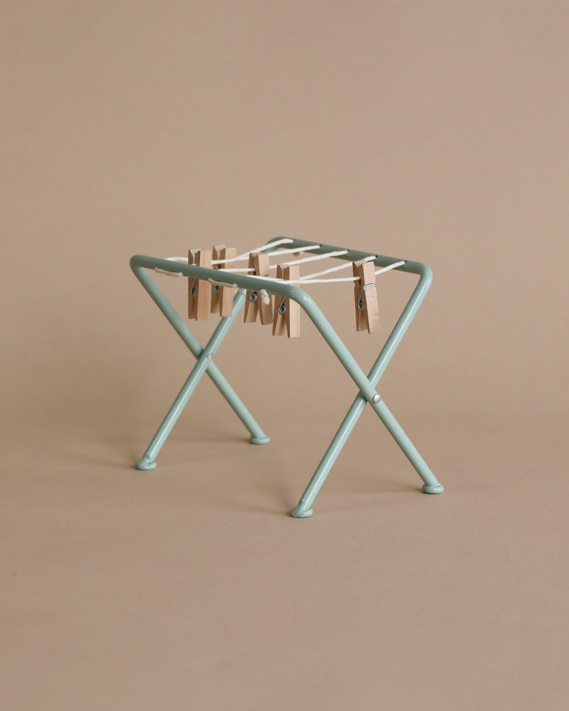 A small green Maileg Miniature Drying Rack with a string tied across its top, holding several wooden clothespins, is displayed against a plain beige background. Ideal for dollhouses or organizing small parts.