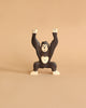 A Holztiger Handmade Wooden Safari Animals Set features a small wooden monkey figurine with raised arms, painted in dark brown and cream, displayed on a beige surface with a plain beige background.