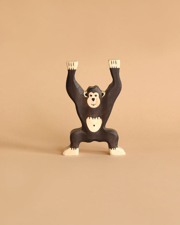 A wooden toy chimpanzee with outstretched arms stands on a plain beige background. Handcrafted from maple and beech wood, this Holztiger Chimpanzee, Standing figure is designed with simplified features and painted in dark brown with white accents on its hands, feet, and face.