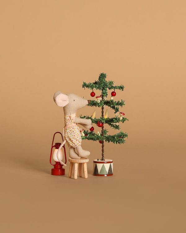 A Maileg mouse sits on a Maileg table and stool, decorating a miniature Christmas tree with red ornaments from the Maileg Holiday Tree Decorating Set. A tiny red lantern completes the festive scene against a beige background.
