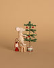 A Maileg mouse sits on a Maileg table and stool, decorating a miniature Christmas tree with red ornaments from the Maileg Holiday Tree Decorating Set. A tiny red lantern completes the festive scene against a beige background.