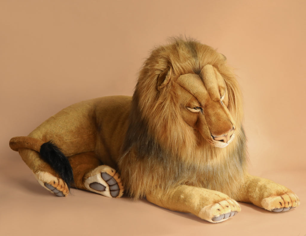 The Extra Large Lion, 39'' – FINAL SALE, is a realistic plush with hand-sewn details and a full mane. It lies on its stomach with stretched-out front paws and half-closed eyes against a plain beige background.