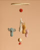 The Maileg Lullaby Friends Mobile hangs against a beige background, showcasing soft fabrics and unique prints: a blue elephant, spotted giraffe, yellow bunny, and pink bear suspended from strings with decorative balls above and below.
