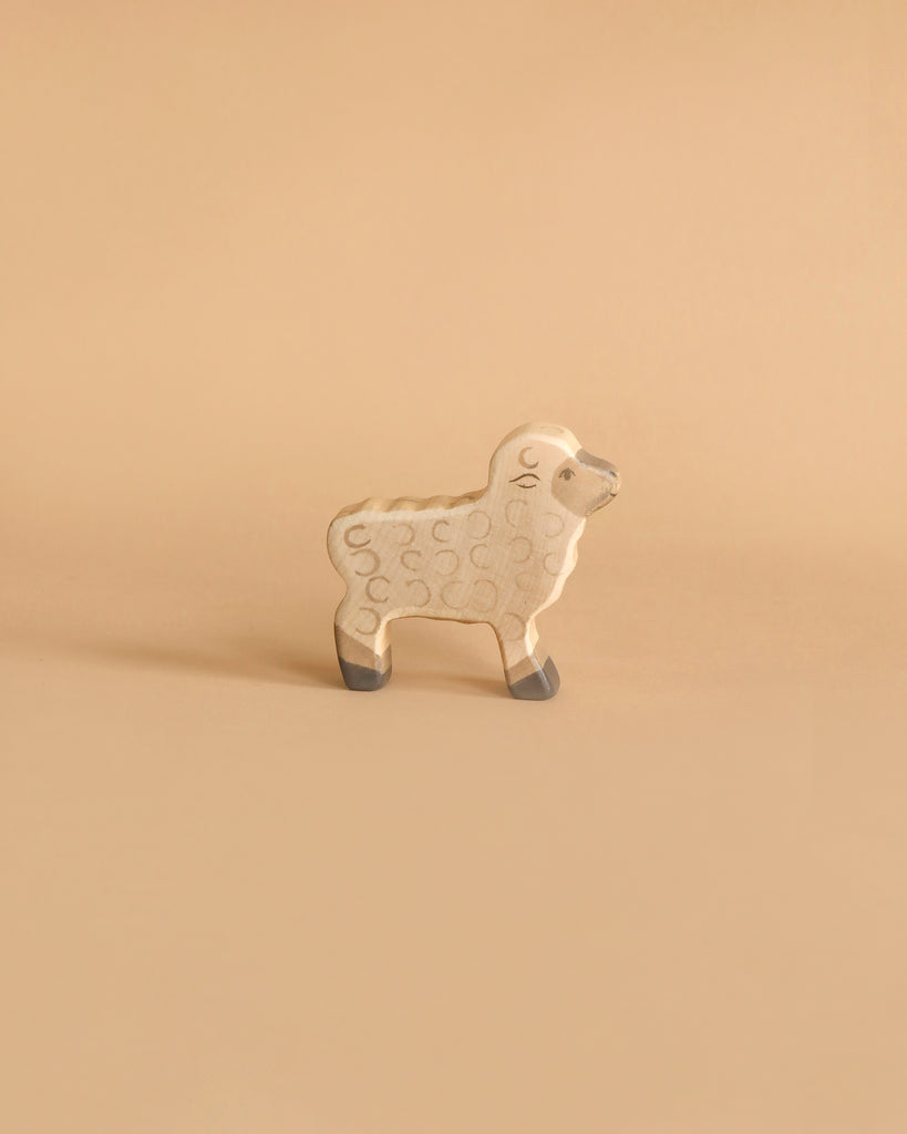 A small wooden sheep from the Holztiger Handmade Wooden Farm Animals Set features a light beige body with subtle swirls and gray accents on its face and feet, styled like classic Holztiger figures, displayed on a plain beige background.