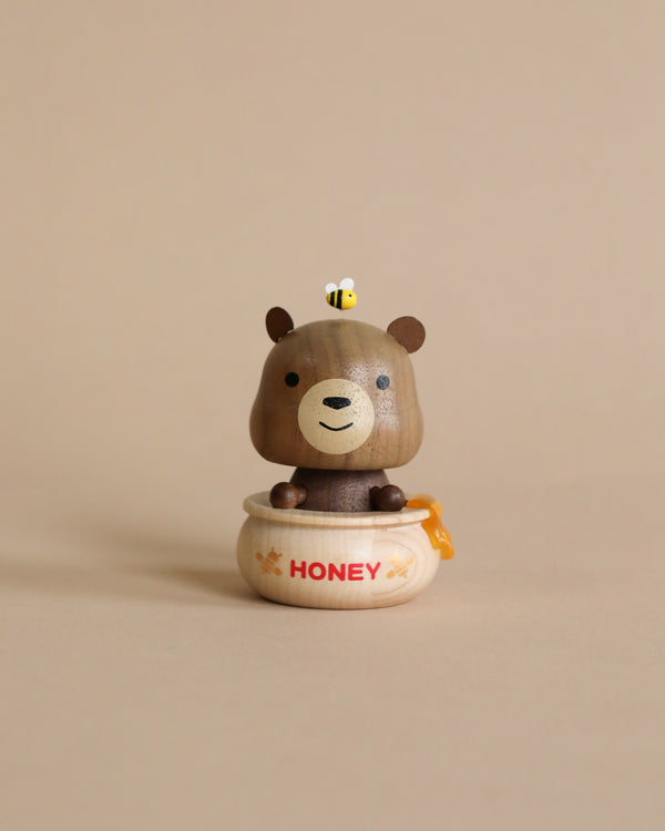 A small Wooden Honey Bear Bobblehead figurine of a smiling cartoon bear sitting inside a honey pot, with a tiny bee on its head, against a beige background.