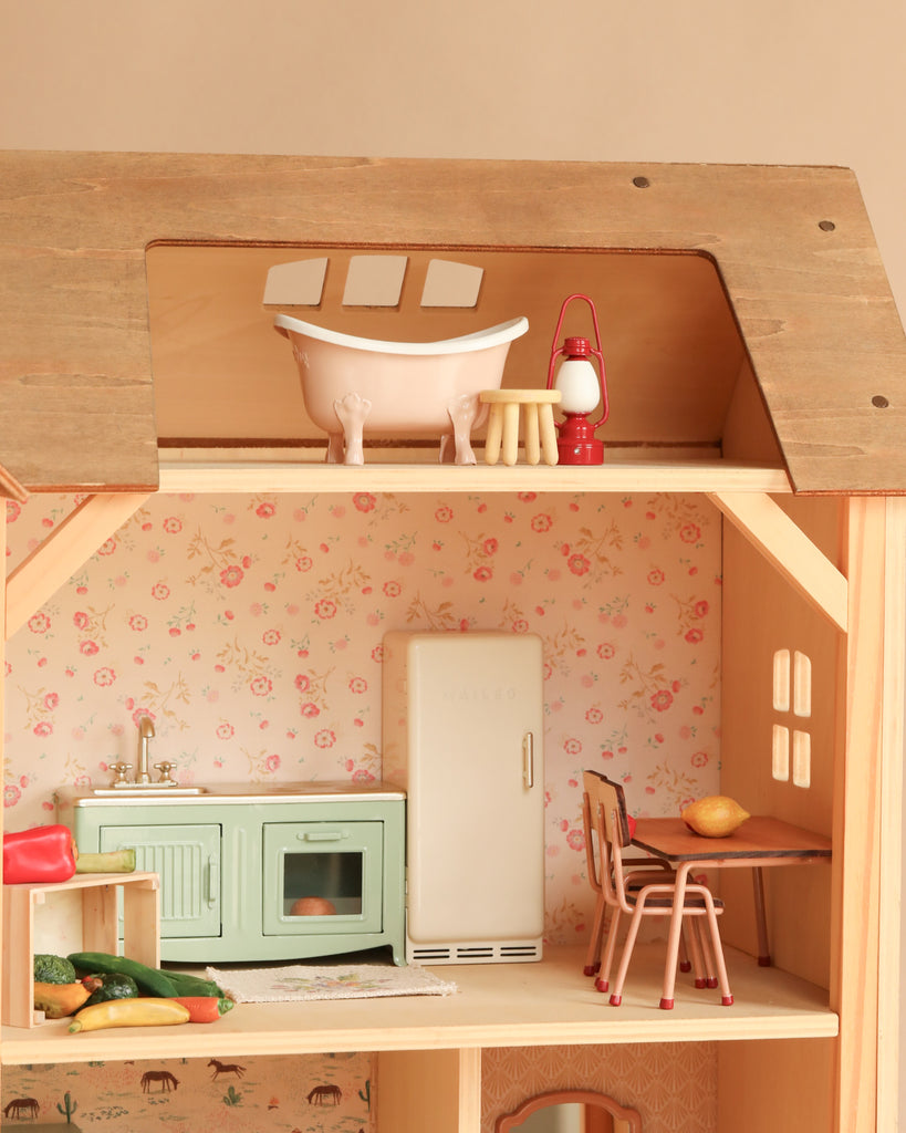 Introducing the Maileg Fully Furnished Farmhouse Set + Surprise Gift: a charming wooden dollhouse complete with a kitchen, dining table, and chairs—perfect for displaying Maileg Mouse Furniture and delightful Dollhouse Accessories. The upper floor features a pink bathtub, stool, and red lantern for imaginative play. In the kitchen you’ll find a green stove, white fridge, and fruit on the table, all set against beautiful floral wallpaper throughout this enchanting farmhouse setting.
