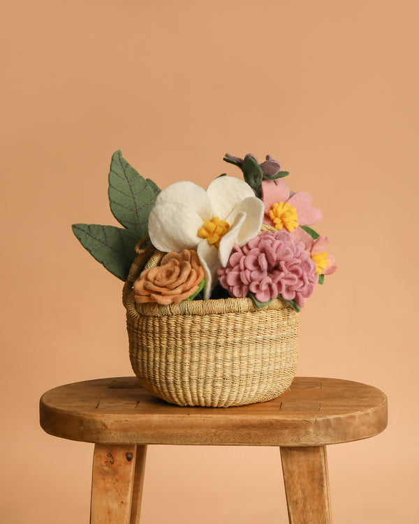 The Basket of Felt Flowers features a vibrant wool felt bouquet with green leaves, arranged in a woven basket and displayed on a wooden stool against a peach background for an everlasting floral touch.