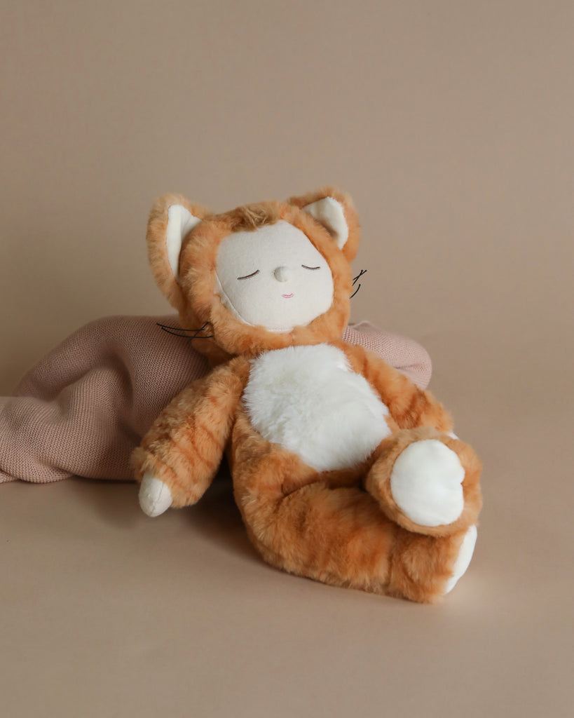 A plush toy resembling a cute, small tabby cat with orange and white fur is lying on its back with its eyes closed, appearing to be asleep. The background is a soft, neutral beige, with a light pink blanket partially visible behind the Olli Ella Cozy Dinkum Doll - Tabby Cat Jinx (ships in approximately one week).