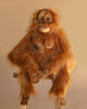 The Hansa Orangutan Monkey Stuffed Animal, featuring lifelike long orange fur, sits on a wooden stool against a plain beige background with its hands resting on its knees.