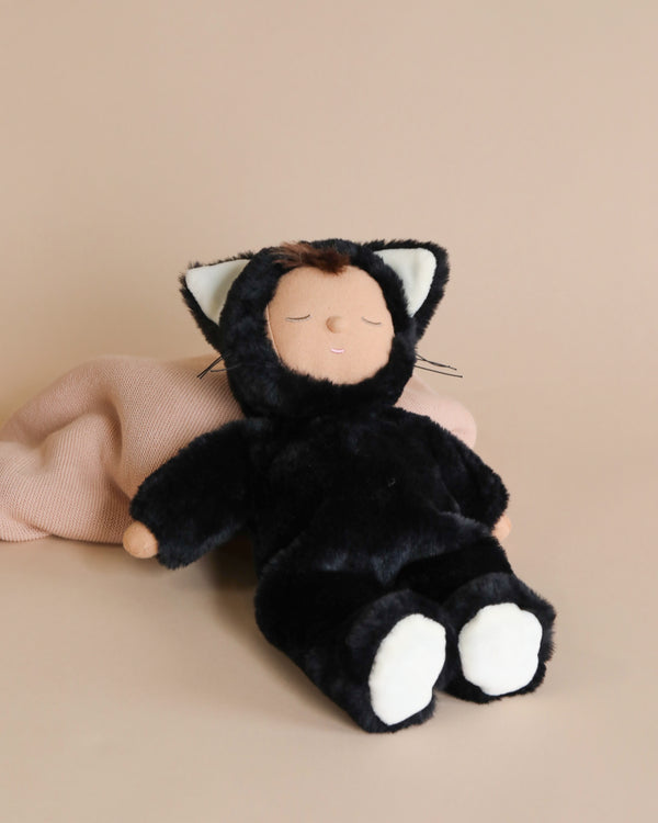 A plush toy resembling a small child dressed in a black and white cat costume, sitting against a soft beige background. The toy, part of the Olli Ella Cozy Dinkum Doll series named Black Cat Nox.