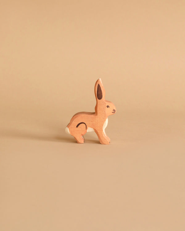 A small wooden Holztiger Rabbit, Standing stands against a plain beige background. Handcrafted out of maple and beech wood, the rabbit is depicted in a sitting position with its ears upright and alert. With a natural wood finish, it features minimal painted details for the eyes, nose, and inner ears.