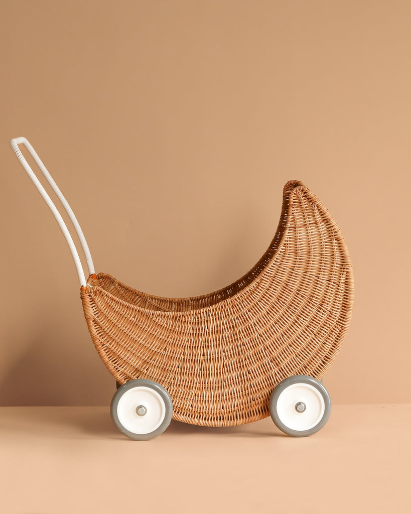 The Olli Ella Moon Strolley is a crescent moon-shaped wicker basket with white wheels and a handle, crafted from natural rattan. Set against a beige background, it’s perfect for imaginative play.