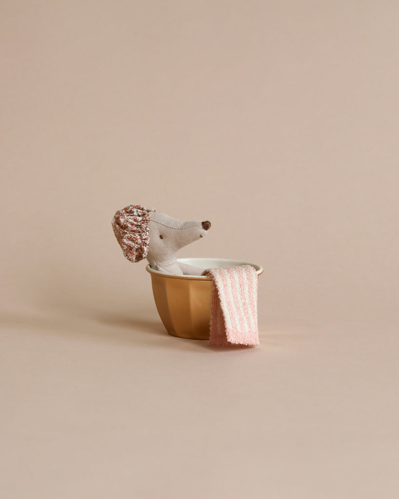 The Maileg Little House Bathroom Set features a small stuffed dog with a textured hat in a gold bowl, next to miniature mouse furniture and a pink-striped towel, all set against a beige background.