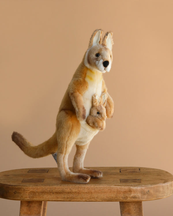 The Kangaroo Mom Stuffed Animal, featuring a baby joey in its pouch and crafted in the style of HANSA animals, stands on a wooden stool against a plain beige background.