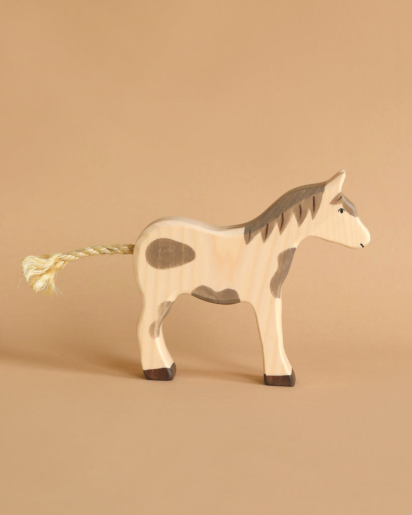 A handcrafted wooden Holztiger Horse, Mustang toy with painted gray spots stands against a plain beige background. This HOLZTIGER figure boasts a braided rope tail and detailed facial features, reflecting the exquisite craftsmanship for which these Made in Europe toys are renowned.