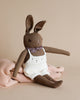 A Polka Dot Club Brown Rabbit in Hand Knit Overalls plush toy with long ears, wearing a white knitted dress and a floral scarf, sitting against a light brown backdrop.