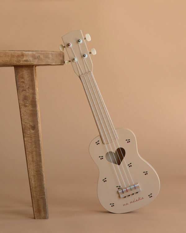 The Wooden Ukulele - Cherry, a charming cream-colored instrument with a heart-shaped sound hole and paw print accents, leans against a wooden stool on a beige background—an adorable, eco-friendly choice for music lovers.