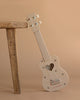 The Wooden Ukulele - Cherry, a charming cream-colored instrument with a heart-shaped sound hole and paw print accents, leans against a wooden stool on a beige background—an adorable, eco-friendly choice for music lovers.