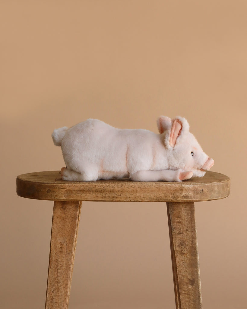 The Pig Stuffed Animal 11" lies on its side on a simple wooden stool, set against a plain beige background, showcasing its realistic plush design.