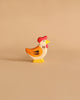 A Holztiger Handmade Wooden Farm Animals Set toy hen, painted yellow, orange, and white with a red comb, is displayed on a plain beige background.