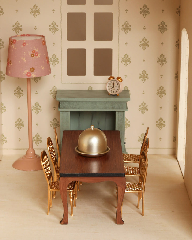 Create a charming dining room in your Maileg dollhouse with the Maileg Fully Furnished Dollhouse Set + Gift With Purchase, which includes a wooden table, six gold chairs, silver cloche, floral lamp, green mantel with clock, and stylish wallpaper.